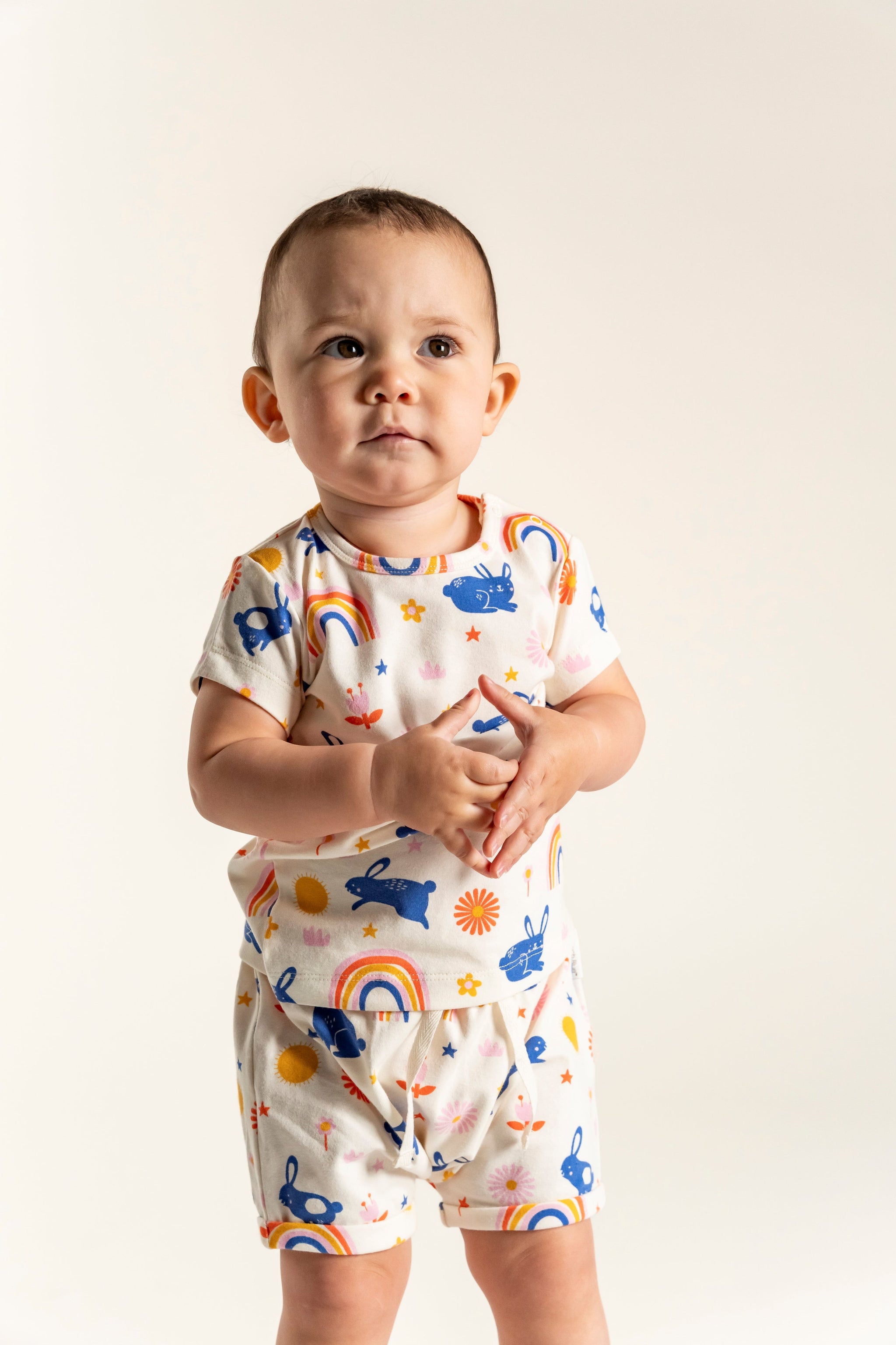 Toddler girl wearing a short sleeve t-shirt with a colorful bunnies and rainbow print, featuring suns, flowers, and stars. Made from soft organic cotton, perfect for casual wear and playtime.