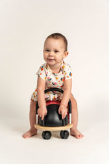 Smiling toddler girl wearing a short sleeve t-shirt with a bright sunny flower print, sitting on a ride-on toy. Made from soft organic cotton, ideal for casual wear, playtime, and outdoor fun.