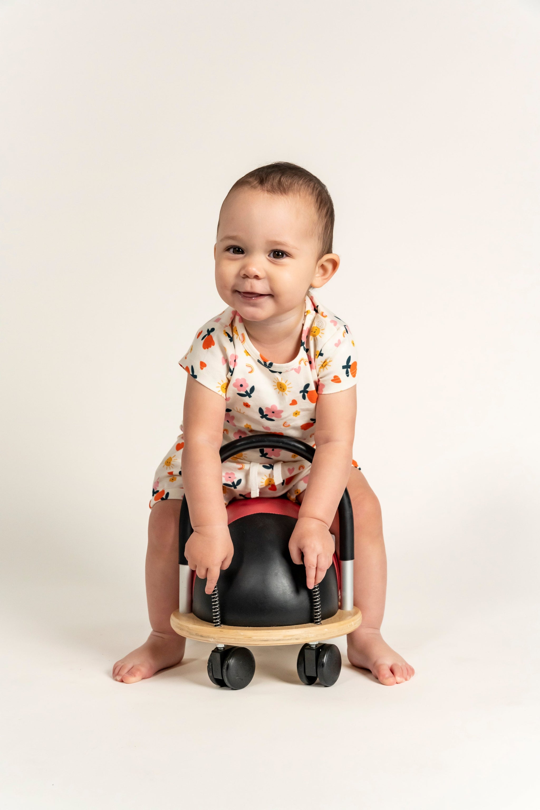 Smiling toddler girl wearing a short sleeve t-shirt with a bright sunny flower print, sitting on a ride-on toy. Made from soft organic cotton, ideal for casual wear, playtime, and outdoor fun.