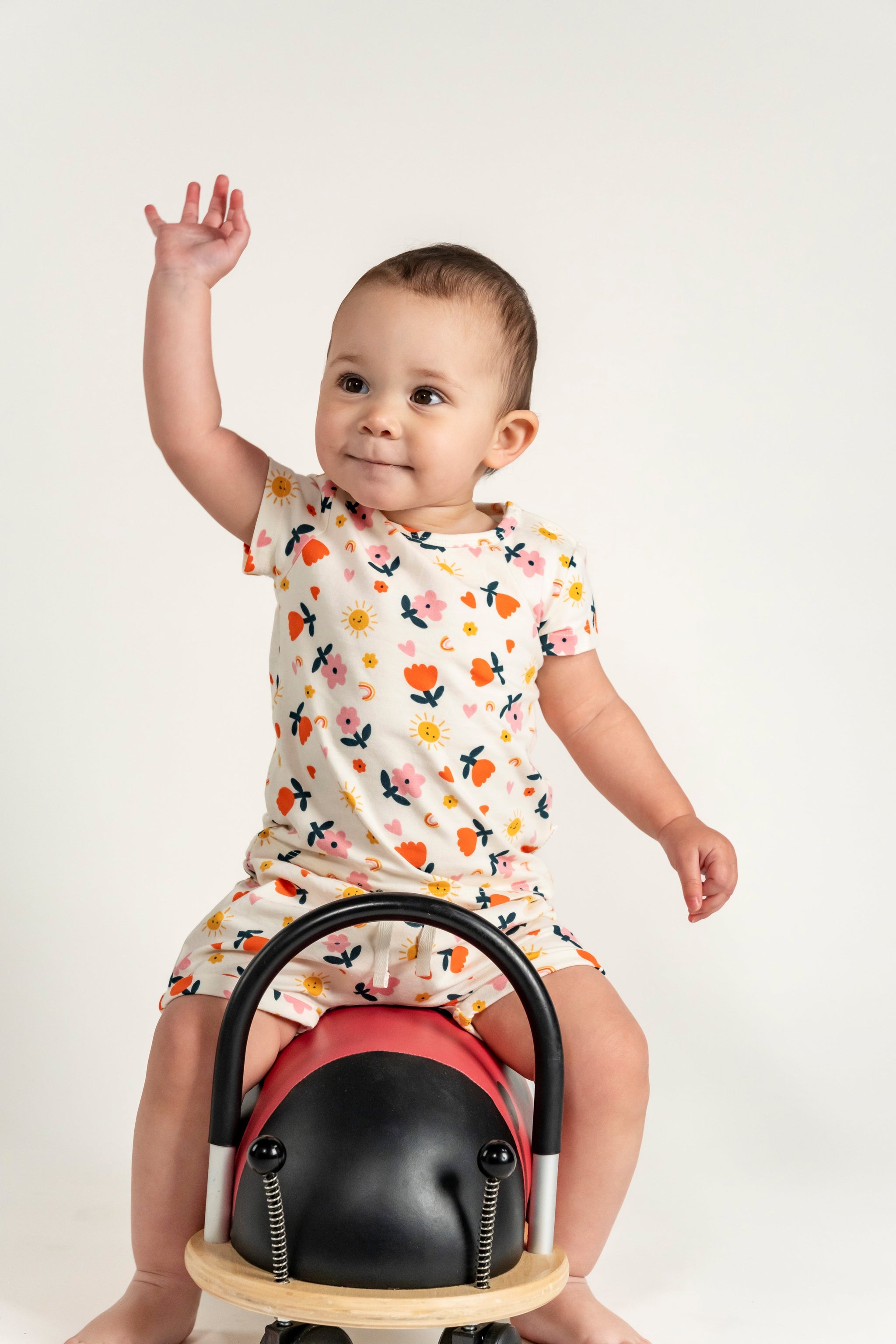 Smiling toddler girl sitting on a ride-on toy while wearing a short sleeve t-shirt with a colorful sunny flower print. Made from soft organic cotton, ideal for casual wear, playtime, and outdoor fun.