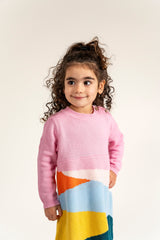 Toddler wearing Serenity Skies Cotton Knitted Sweater Dress featuring pink sleeves and colorful geometric patterns in yellow, blue, orange, and green. Cozy, soft cotton perfect for casual fall/winter wear.