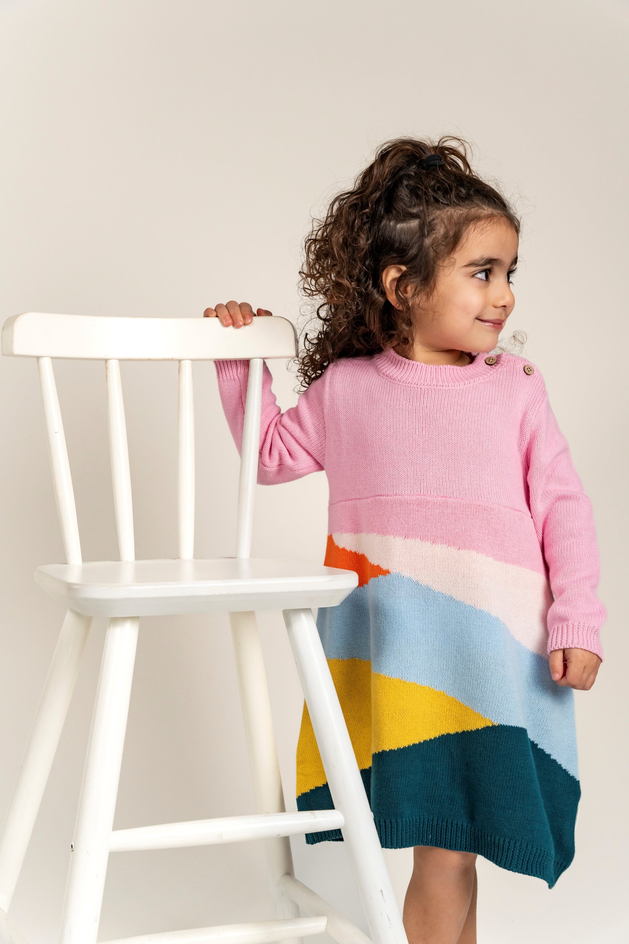 Toddler wearing Serenity Skies Cotton Knitted Sweater Dress featuring pink sleeves and colorful geometric patterns in yellow, blue, orange, and green. Soft and cozy, perfect for fall/winter casual wear.