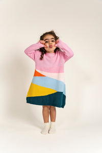 Toddler wearing Serenity Skies Cotton Knitted Sweater Dress with pink sleeves and vibrant geometric patterns in yellow, blue, orange, and green. Soft, cozy cotton perfect for playful fall/winter outfits.