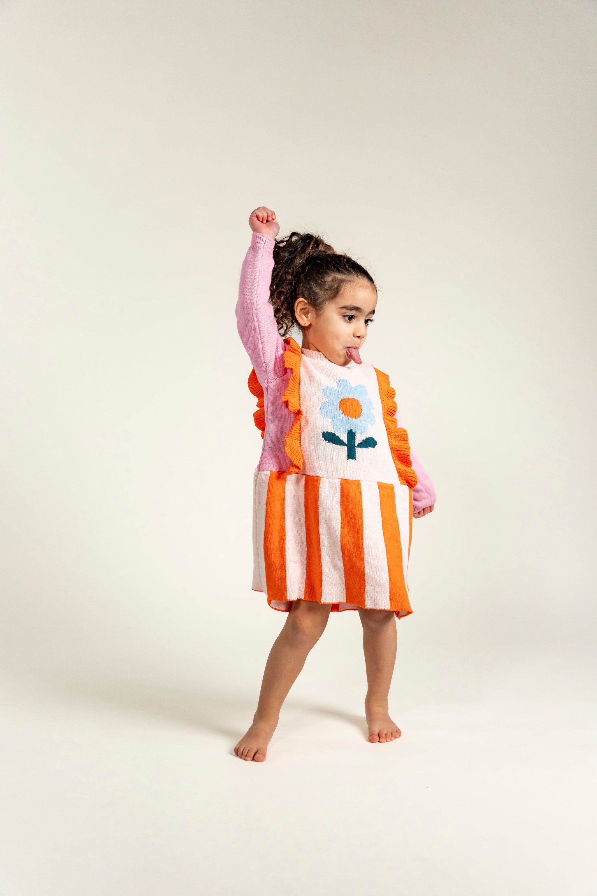 Toddler wearing Petal Playtime Cotton Knitted Dress with pink sleeves, orange ruffles, stripes, and a blue flower design. Soft, cozy, long-sleeved dress perfect for playful fall/winter casual outfits.