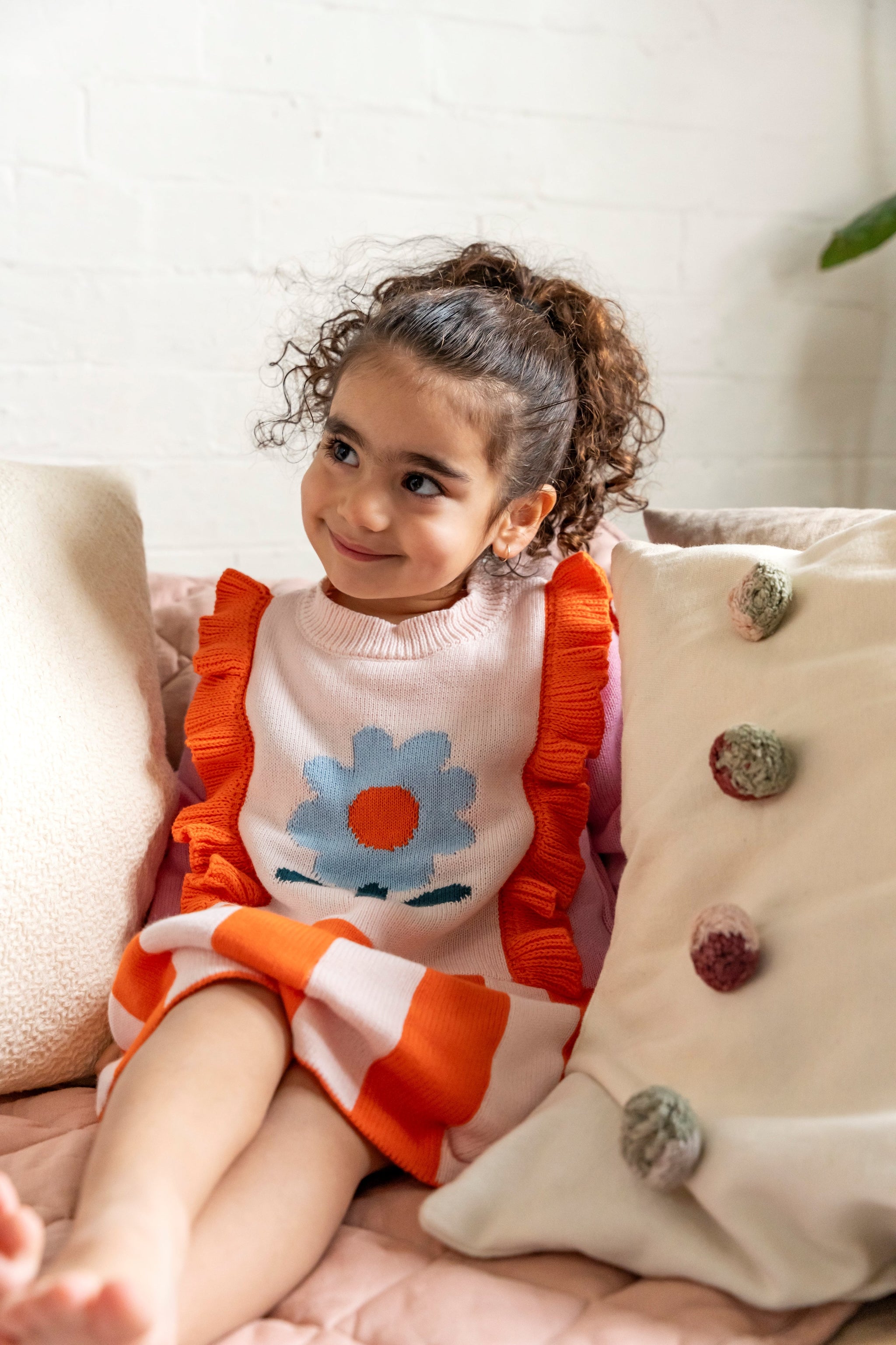 Toddler sitting on a couch wearing the Petal Playtime Cotton Knitted Dress with pink sleeves, orange ruffles, and a blue flower design. Soft, cozy, perfect for casual fall/winter outfits.