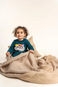 Toddler sitting wrapped in a cozy blanket wearing Pompom Wonderland Cotton Knitted Sweater Dress in dark green with a white cat design and colorful pom-poms. Soft, long-sleeved, perfect for fall/winter wear.