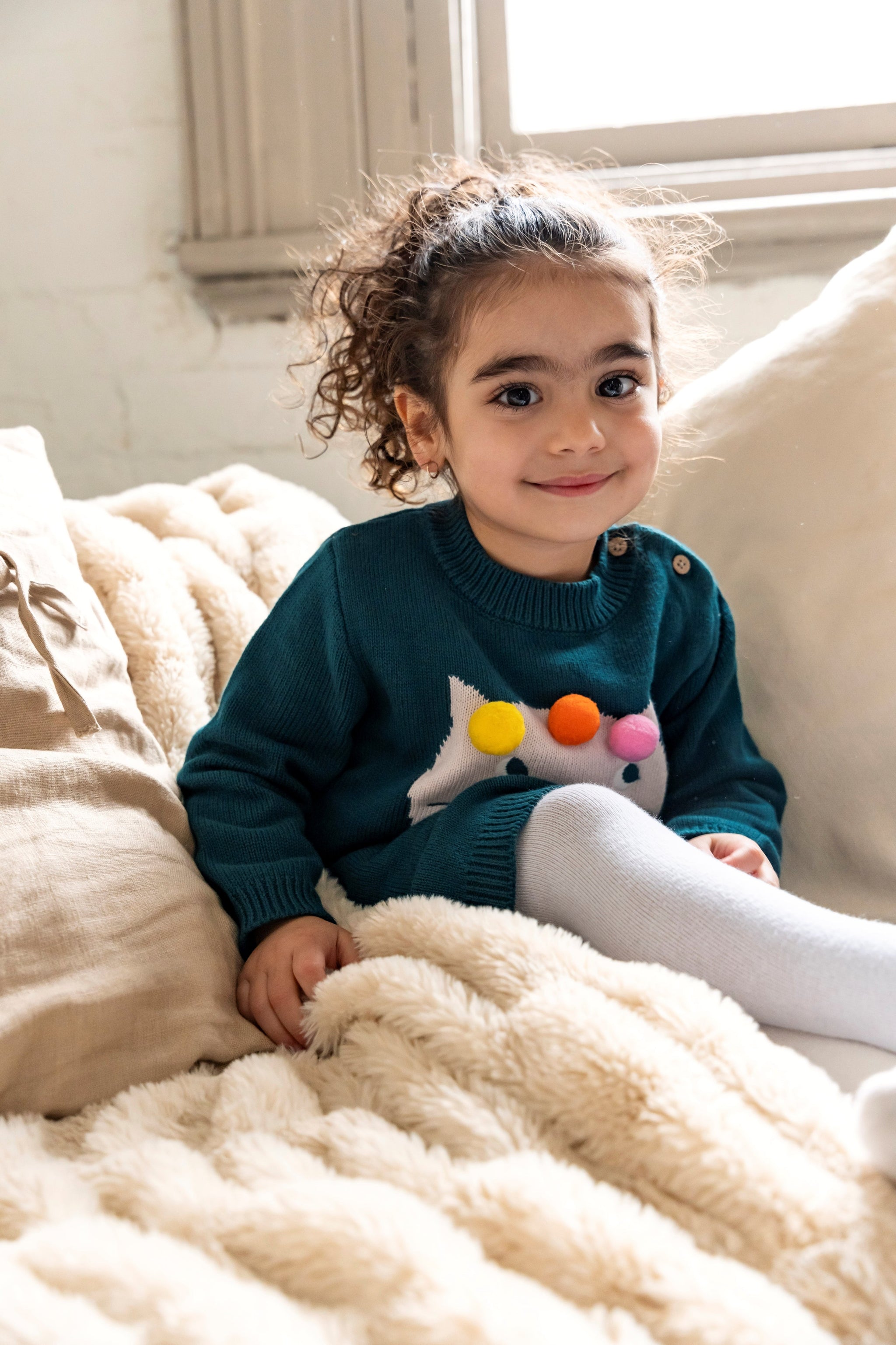 Toddler sitting on a cozy couch wearing Pompom Wonderland Cotton Knitted Sweater Dress in dark green with a white cat design and colorful pom-poms. Soft, long-sleeved, perfect for fall/winter outfits.