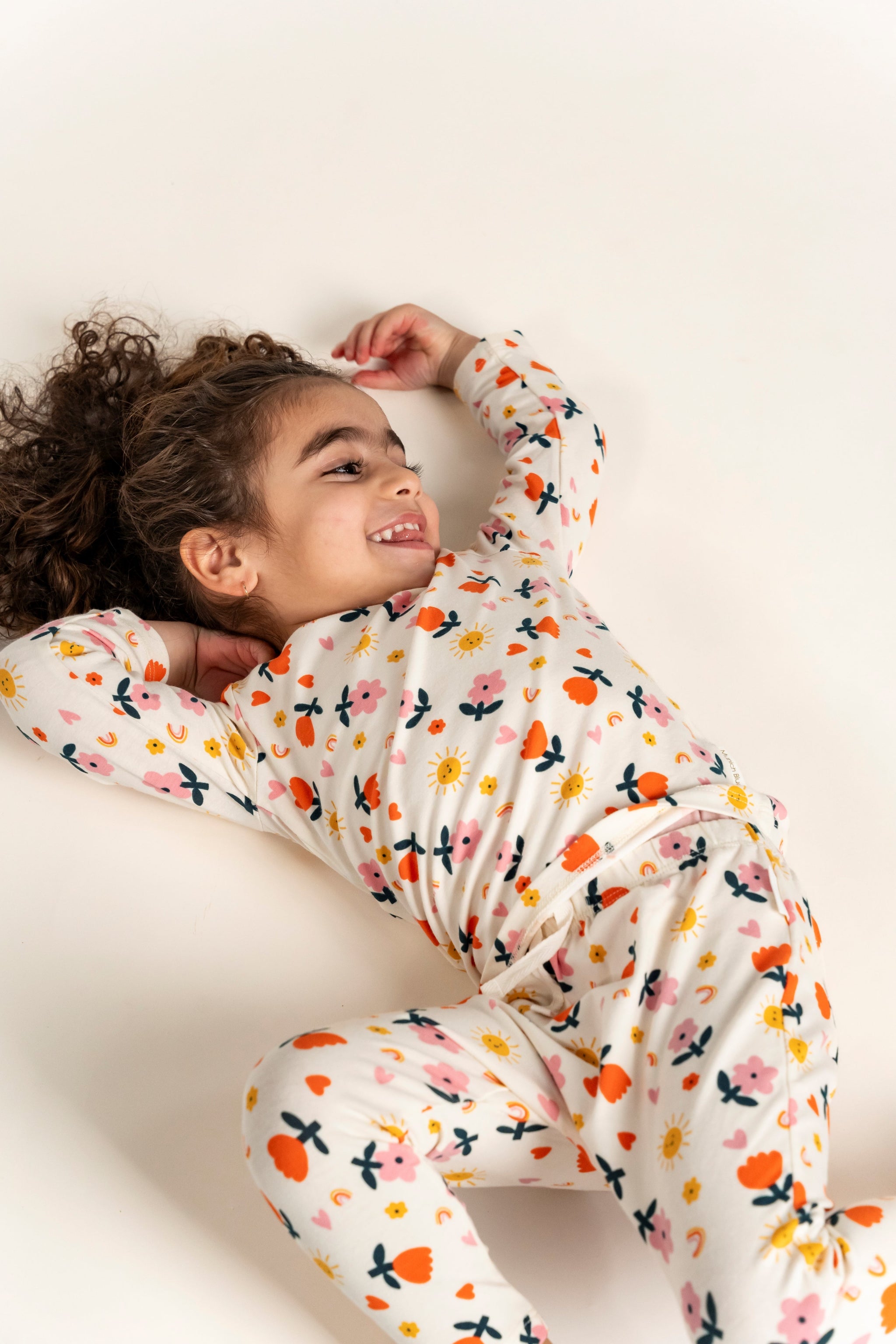 Toddler Girl smiling and lying down, wearing a Long Sleeve T-shirt and pants set with a vibrant Sunny Flower Print. Featuring colorful flowers, hearts, and sun designs, perfect for casual wear.