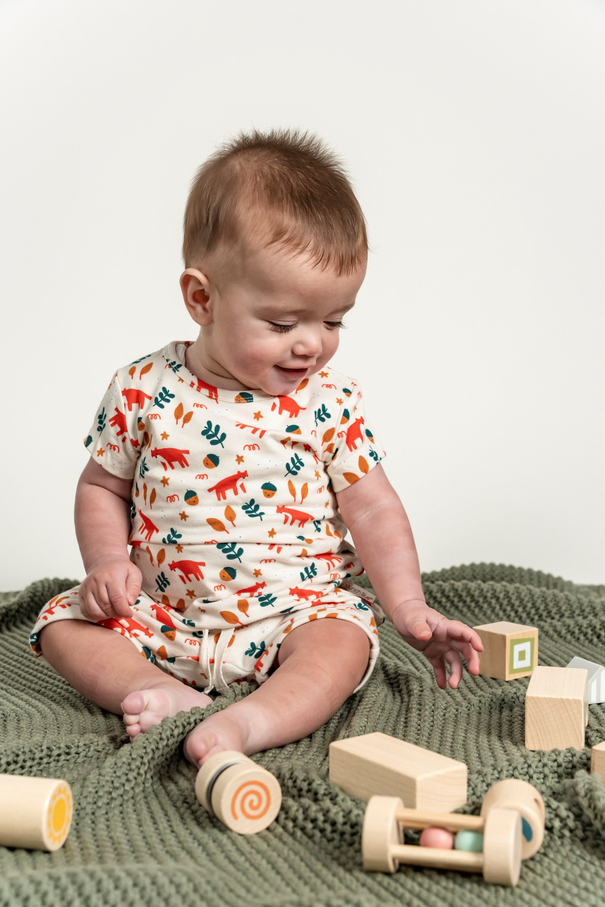 Smiling toddler boy playing with wooden toys while wearing a short sleeve t-shirt with fox forest print, featuring red foxes and green leaves. Soft organic cotton, ideal for casual and everyday wear.