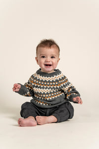 Baby wearing a cozy Fair Isle Sweater Jumper in dark grey with white and orange geometric patterns. Soft cotton, long-sleeved sweater perfect for keeping babies warm during fall and winter.