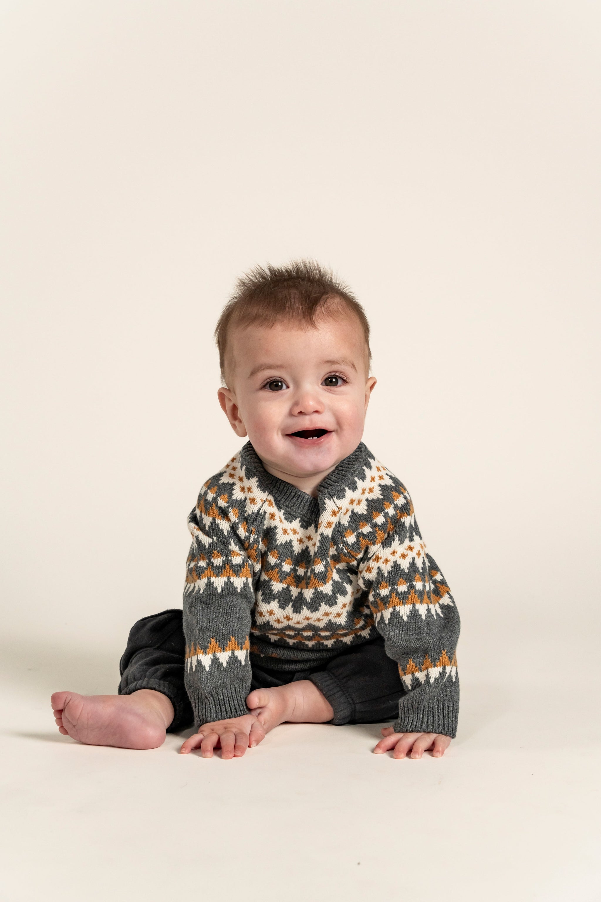 Adorable baby wearing a cozy Fair Isle Sweater Jumper featuring grey, white, and orange geometric patterns. Perfect for keeping little ones warm and stylish during cooler months. 100% soft cotton fabric.