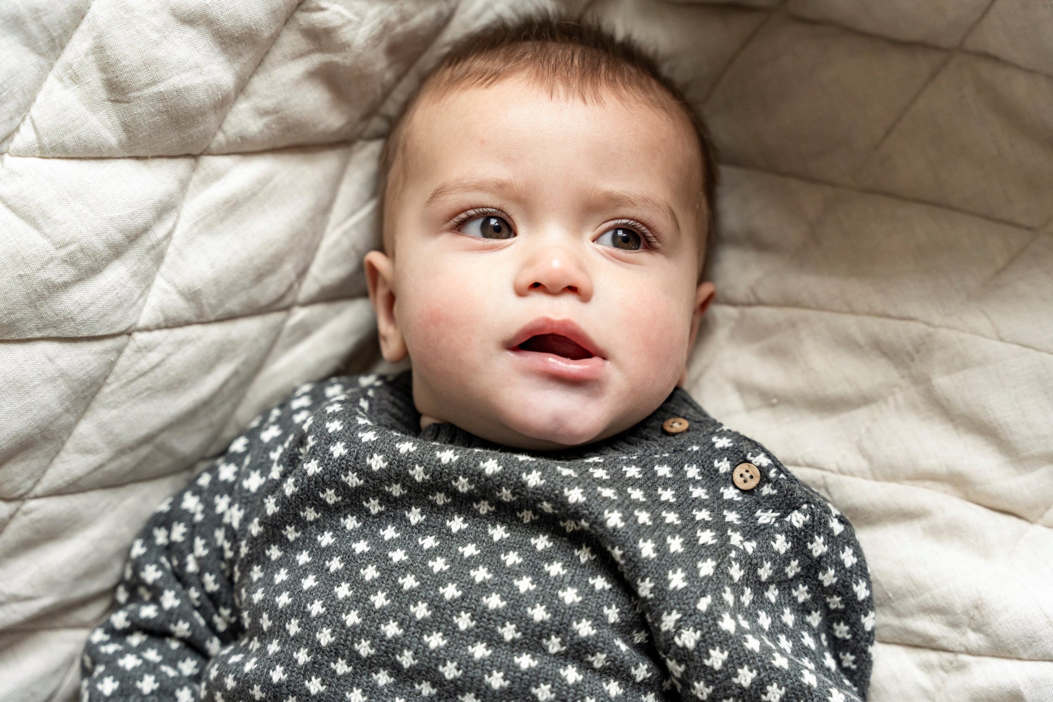 Baby boy laying on a cozy blanket, dressed in a grey Nordic Cotton Knit Jumper with white geometric patterns and wooden button detailing. Perfect for keeping your little one warm and stylish.