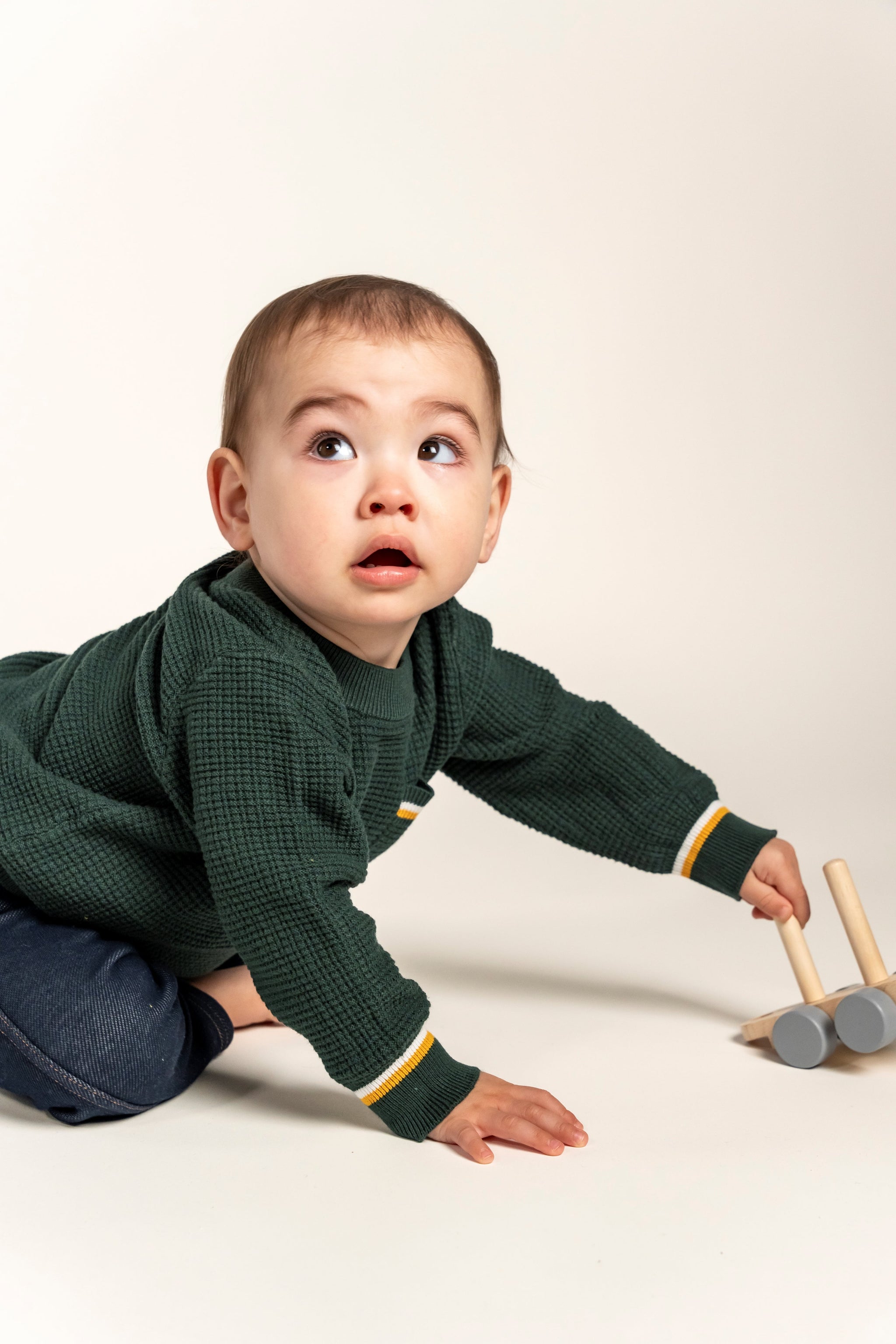 Adorable baby wearing a forest green Waffle Cotton Knit Jumper with yellow and white stripe details on the pocket and cuffs, playing with a wooden toy. Ideal cozy sweater for your little one’s everyday adventures.