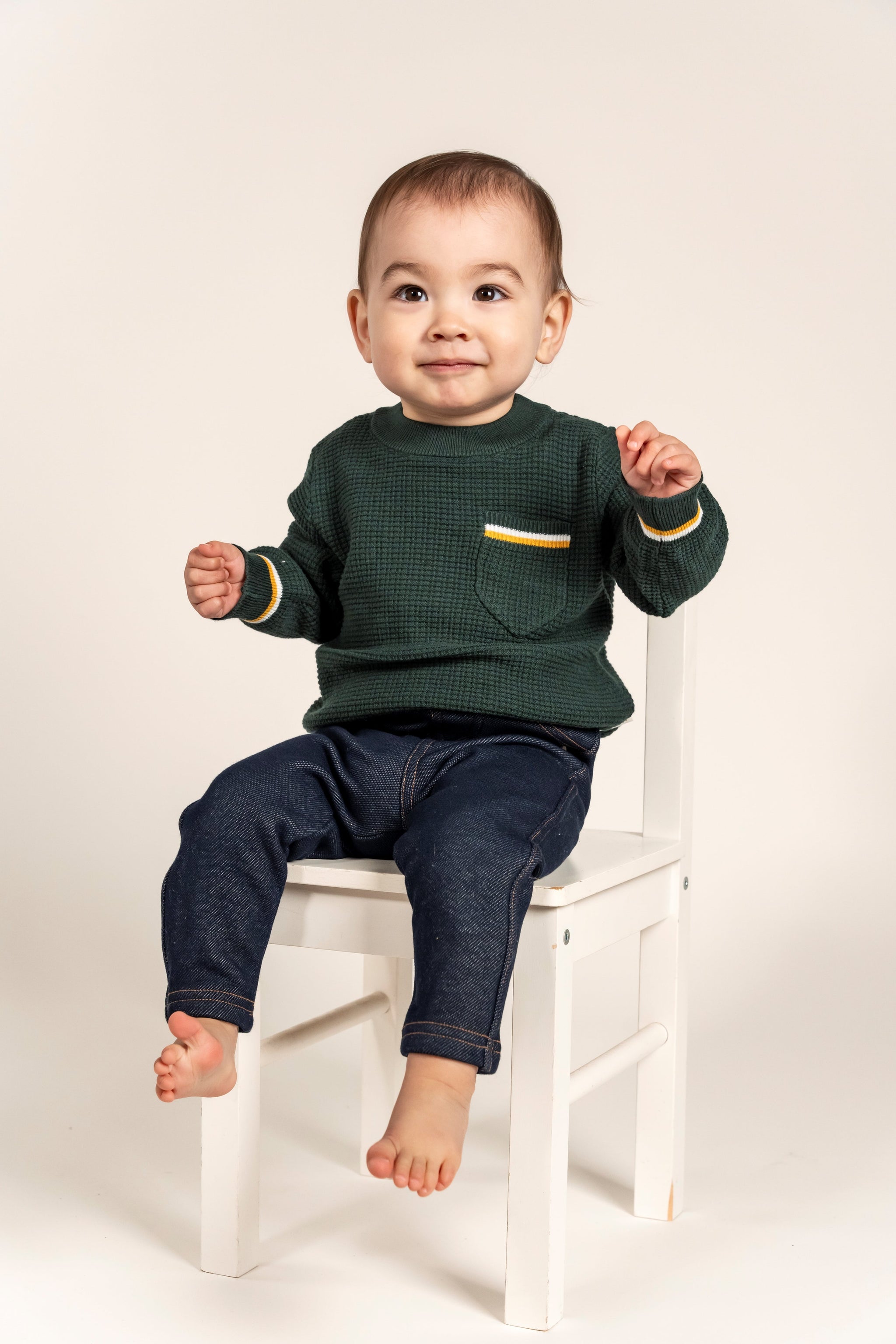 Cute toddler wearing a forest green Waffle Cotton Knit Jumper with yellow and white stripe details on the pocket and cuffs, seated on a white chair. Cozy and stylish, perfect for everyday wear.