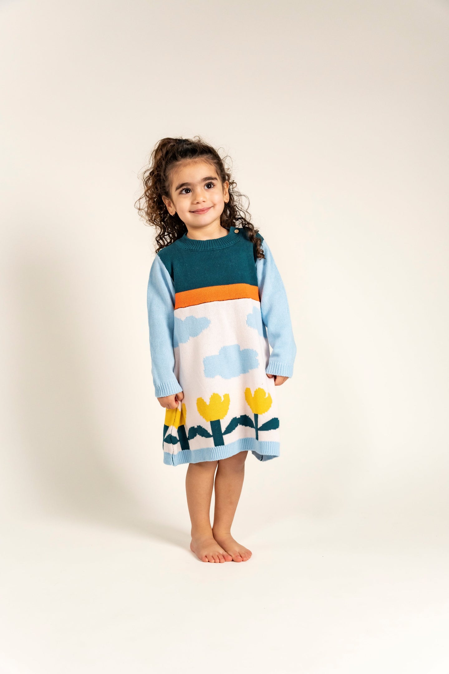 Toddler wearing Cloudy Daydream Cotton Knitted Sweater Dress featuring soft blue sleeves, cloud patterns, yellow tulips, and an orange stripe. Cozy, long-sleeved, ideal for fall/winter casual outfits.