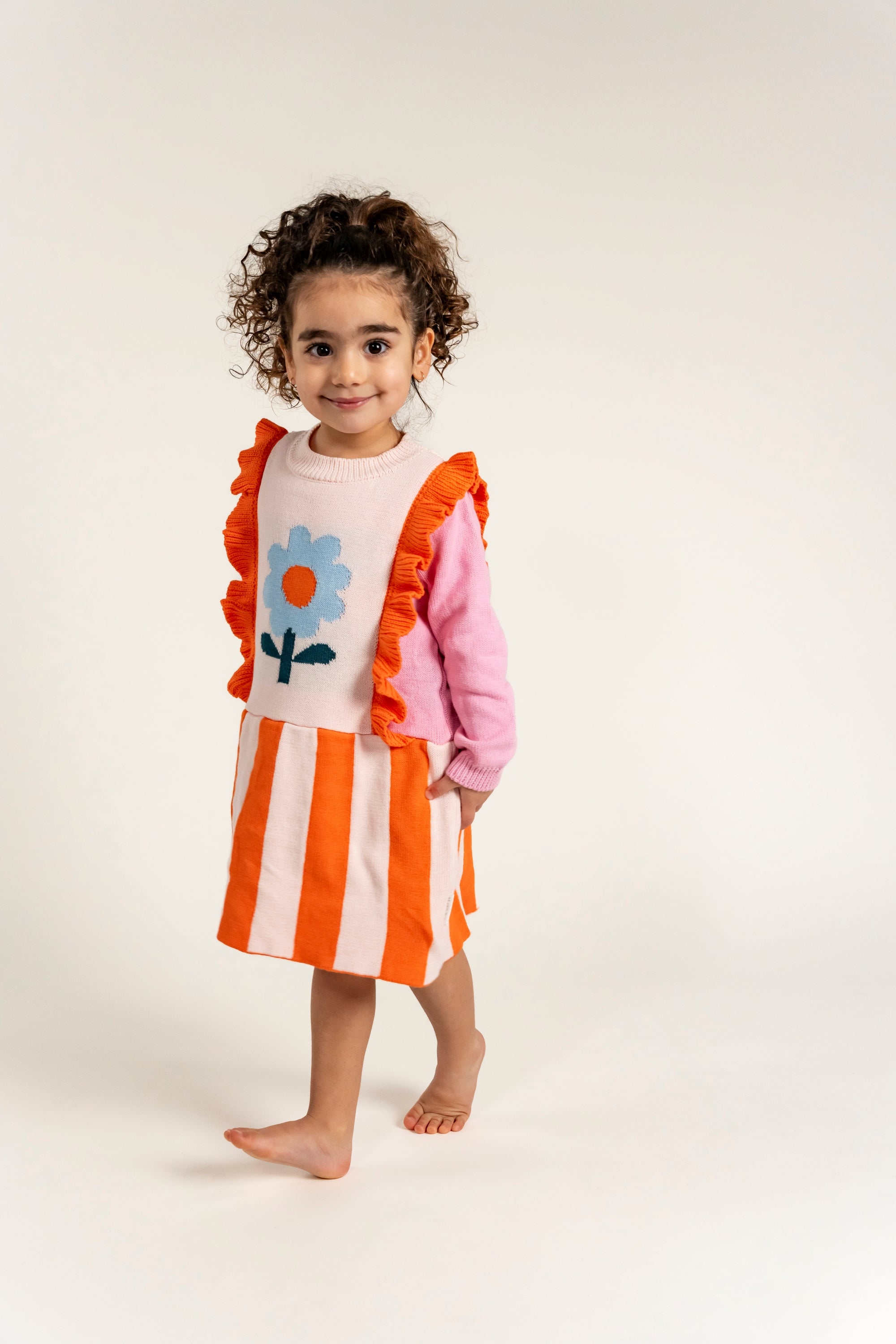 Toddler wearing Petal Playtime Cotton Knitted Dress featuring pink sleeves, orange ruffles, and stripes with a blue flower design. Soft, cozy, long-sleeved dress ideal for fall/winter casual wear.