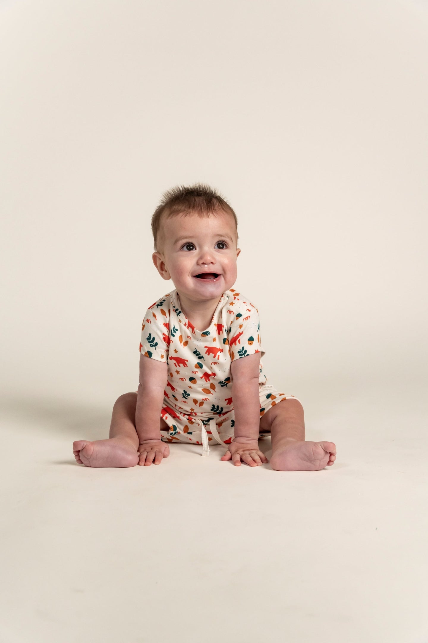 Happy toddler boy sitting and wearing a short sleeve t-shirt with a fox forest print featuring orange foxes and green trees. Soft, organic cotton, perfect for casual wear and outdoor adventures.