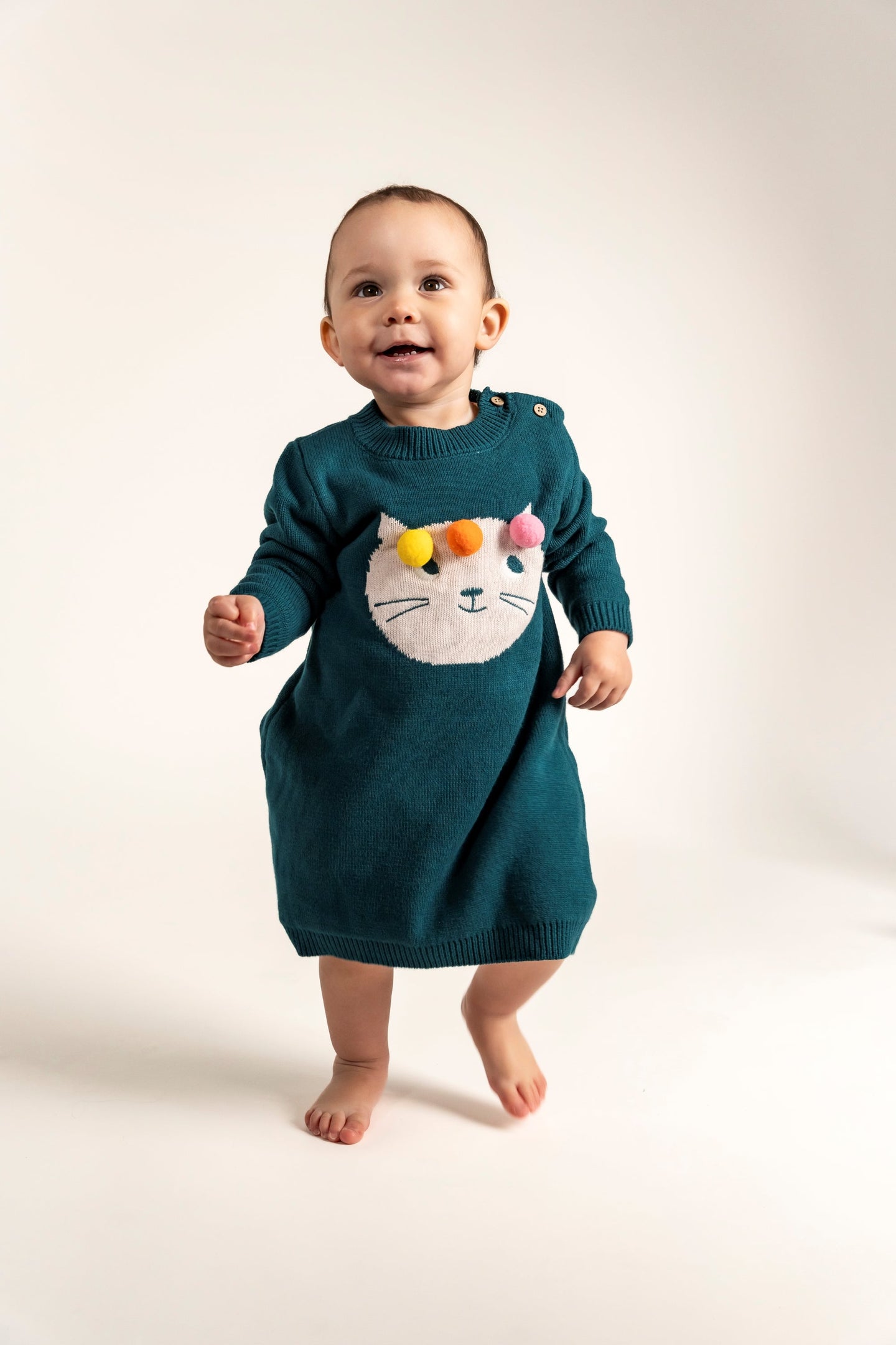 Toddler wearing Pompom Wonderland Cotton Knitted Sweater Dress in dark green with a white cat design and colorful pom-poms. Soft, cozy, long-sleeved, perfect for fall/winter casual outfits for toddlers.