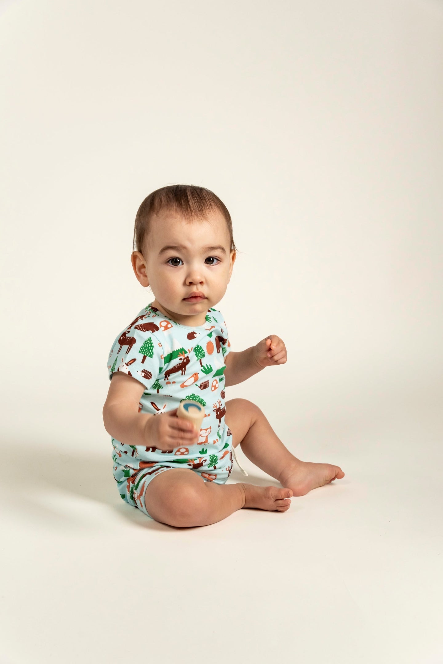 Adorable toddler boy sitting and wearing a short sleeve t-shirt with a forest animal print. The t-shirt features cute illustrations of trees, foxes, and woodland animals. Ideal for everyday wear.