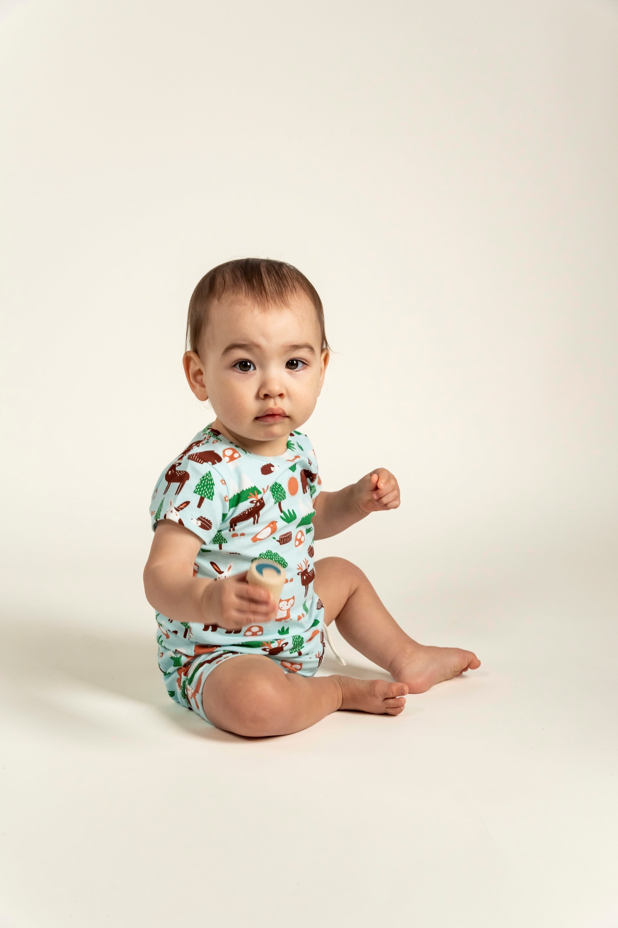 Adorable toddler boy sitting and wearing a short sleeve t-shirt with a forest animal print. The t-shirt features cute illustrations of trees, foxes, and woodland animals. Ideal for everyday wear.