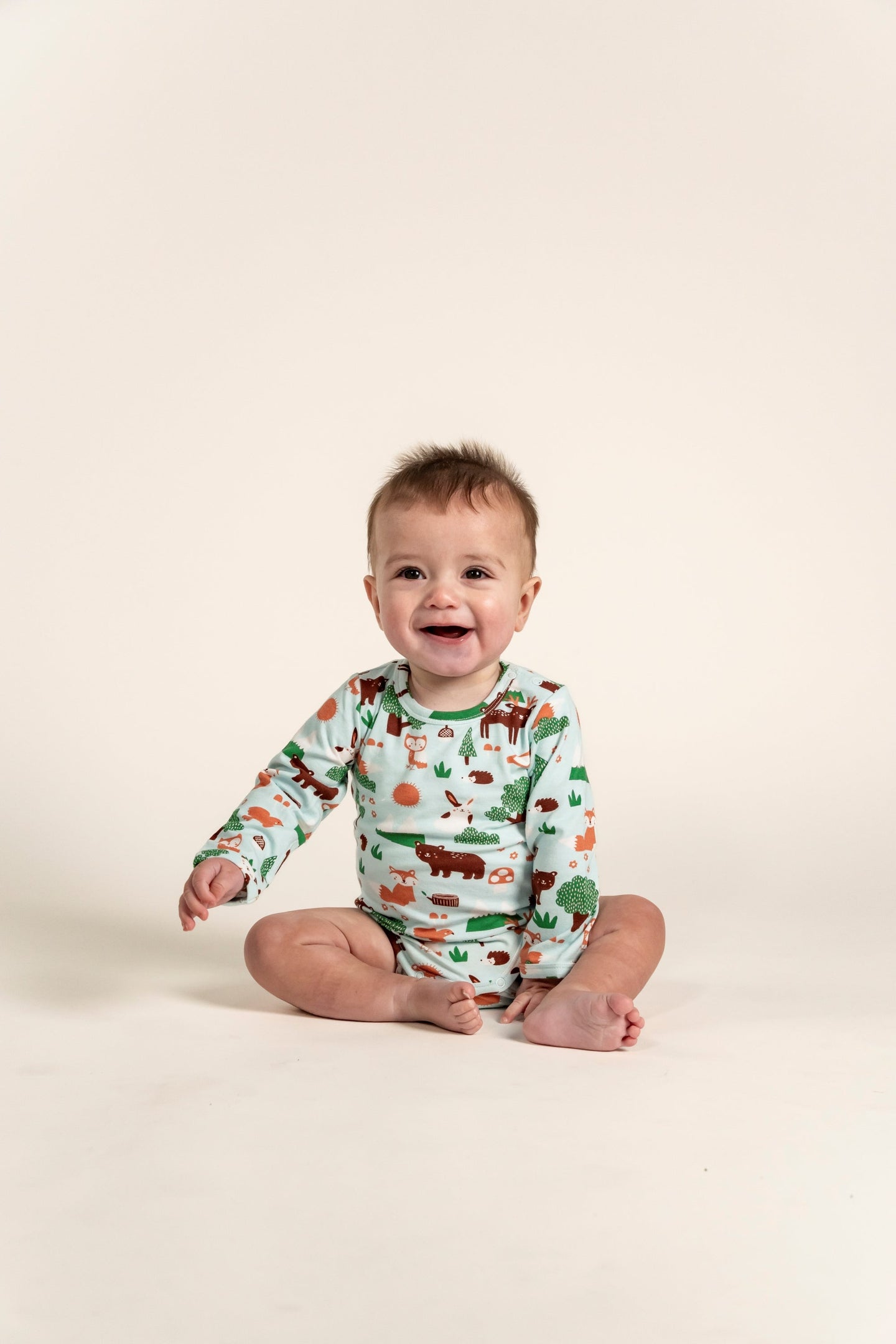 Toddler Boy smiling and sitting while wearing a Long Sleeve Bodysuit with a playful Forest Animal Print. Features woodland creatures like bears, foxes, and trees, perfect for cozy everyday wear.